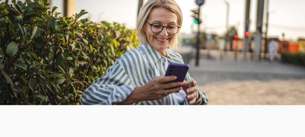 Ältere Frau mit einem gestreiften Hemd sitzt lachend auf einer Bank und hält ein Handy in der Hand